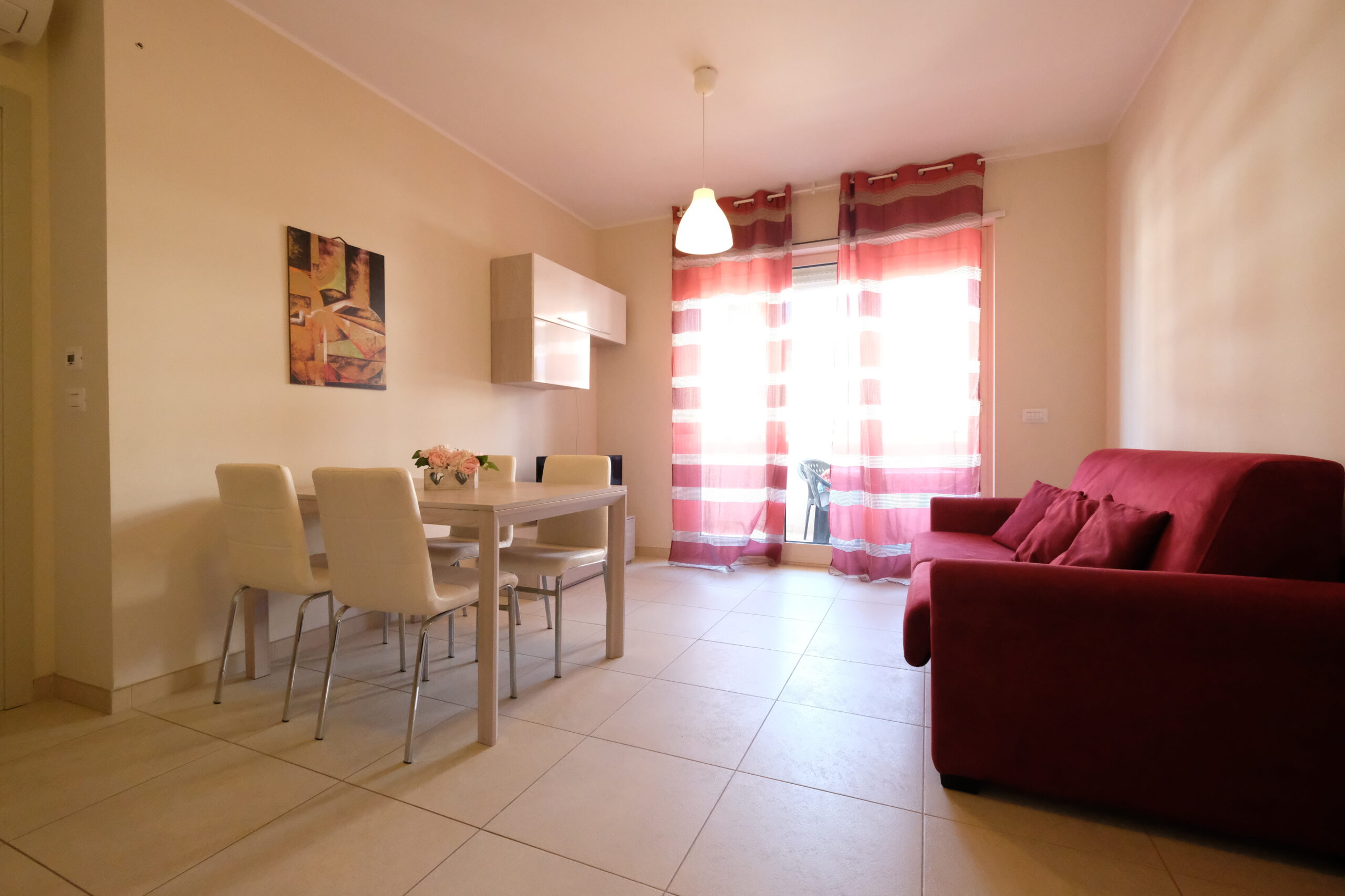 Sala con tavolo e sedie bianche, divano bordeaux e finestra a porta con tende Appartamento Pineto Vacanza Residence Claudia int. F