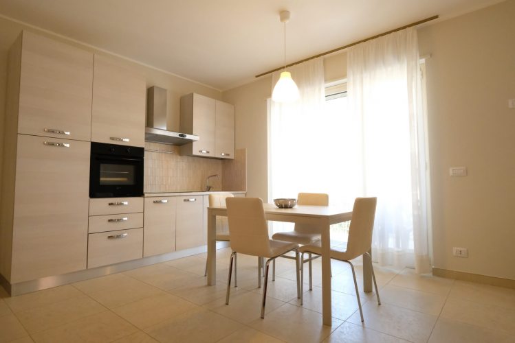 Cucina beige con finestra e tenda e tavolo e sedie bianche Appartamento Pineto Vacanza Residence Claudia int. M